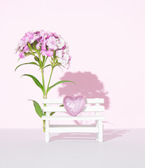 Minimal creative romantic concept of spring flowers, park or garden enjoyment. White bench and Sweet William flowers in full bloom. Pastel pink colored background. Frontal view. Copy space.