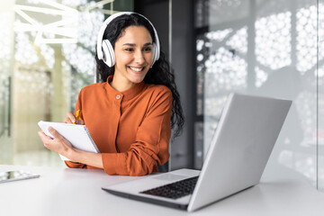 Latin american business woman with curly hair and headphones watching online training course at workplace, woman writing information happy and satisfied with the results of professional development. - obrazy, fototapety, plakaty
