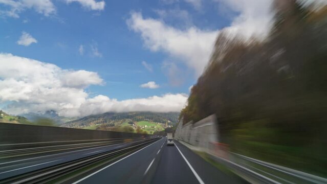 Experience a timelapse POV perspective of highway traffic in Austria as you drive through the picturesque mountain scenery during autumn, surrounded by vivid clouds and blue sky.