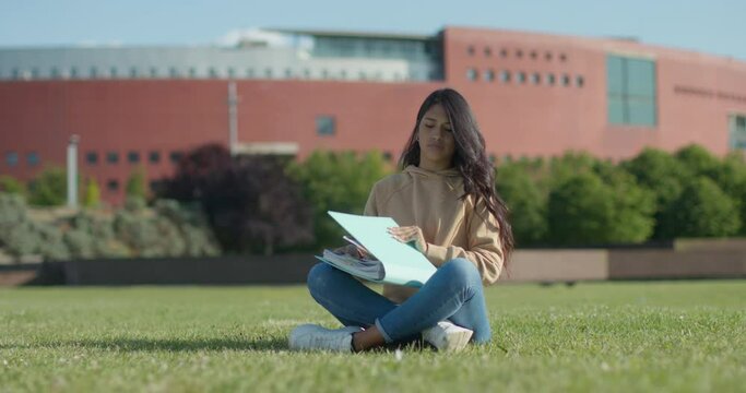Young teenage Latina college student studying for an exam in an outdoor park with books