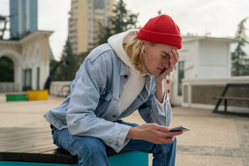 Worried upset millennial guy sitting alone on bench outdoors with smartphone in hands, touching...