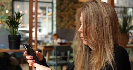 Medium shot young woman disappointed by message on phone in indoor public space. Beautiful genuine stylish warm color interior with plants and big windows in background.