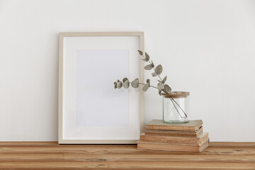  Empty wooden picture frame, poster mockup on white wall background. Glass vase with green eucalyptus tree branches on table and books. Neutral still life interior.Empty copy space.Modern art display