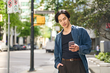 Young smiling asian man holding smartphone using mobile app, reading text message, communication online walking on urban street. Japanese hipster guy shopping online outdoors