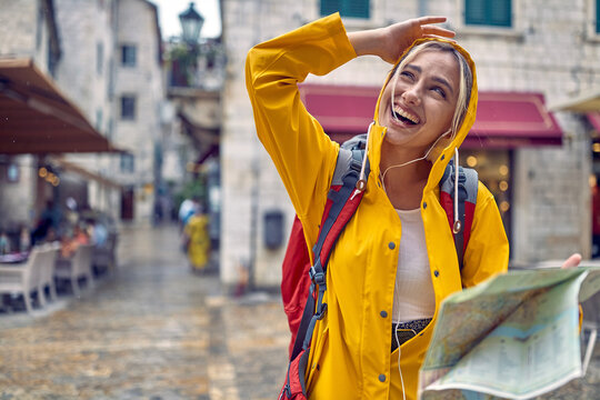 Walk in city in rain. Young tourist woman in raincoat with map, feeling happy, smiling. Travel in new city. Tourism, lifestyle, fun, weather concept.