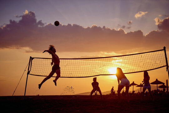 Friends playing volleyball at beach in group, young athletic man jumping hitting ball. Sport, recreation, fun, togetherness, lifestyle concept. - Powered by Adobe
