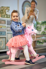 Lovely toddler girl riding a pink unicorn rocking horse in a shop, feeling joyful, mother taking a picture in the background.