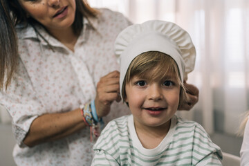 Mother of little blonde boy putting a kitchen hat on his head inside the kitchen.