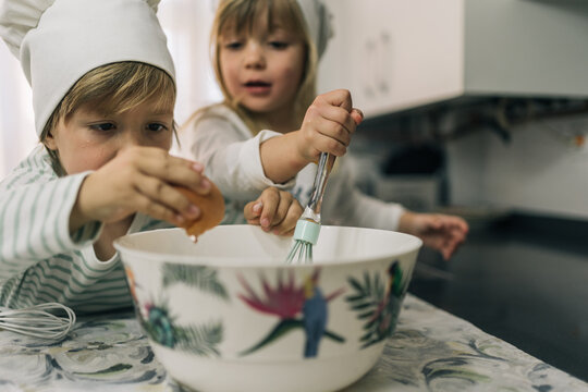 Pair of blond brother and sister children cracking an egg into a bowl to beat it with egg beaters to make a cake.