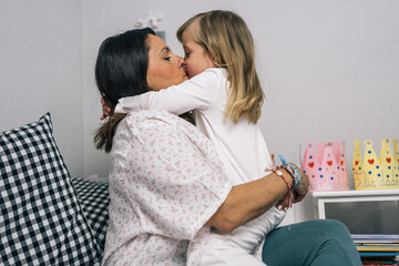 Mother sitting on bed with her blonde daughter giving each other a tight hug and a loving kiss on the mouth.