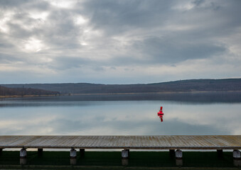 a scenic view of a lake with a wooden dock in the foreground. The sky is cloudy and there are trees in the background. In the water, there is also a red buoy visible.