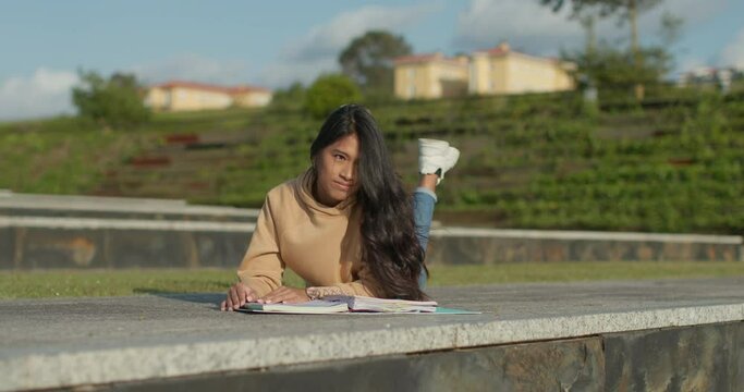 Young teenage Latina college student studying for an exam in an outdoor park with books