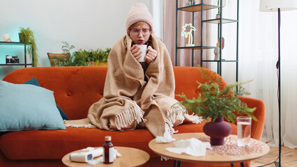 Sick young woman wear hat wrapped in plaid sit alone shivering from cold on sofa drinking hot tea...