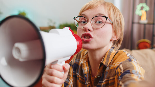 POV selphi portrait of young woman talking with megaphone, proclaiming news, loudly announcing advertisement, warning using loudspeaker to shout speech sitting on sofa. Girl at home room apartment