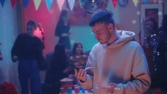 The guy carefully looks at the phone at the party while his friends are having fun and dancing. Reads a message, browses social networks, throws up his hands. Party, celebration, birthday, disco.