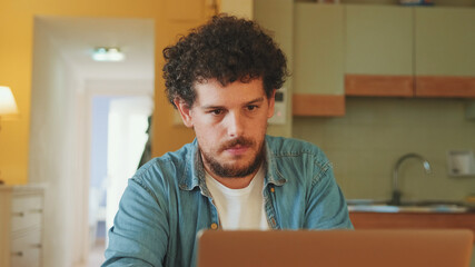 Guy dressed in denim shirt works on laptop while sitting in cozy kitchen, freelancer working