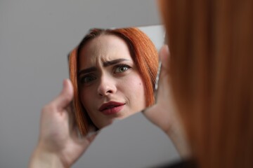 Young woman looking at herself in shard of broken mirror on light grey background, closeup
