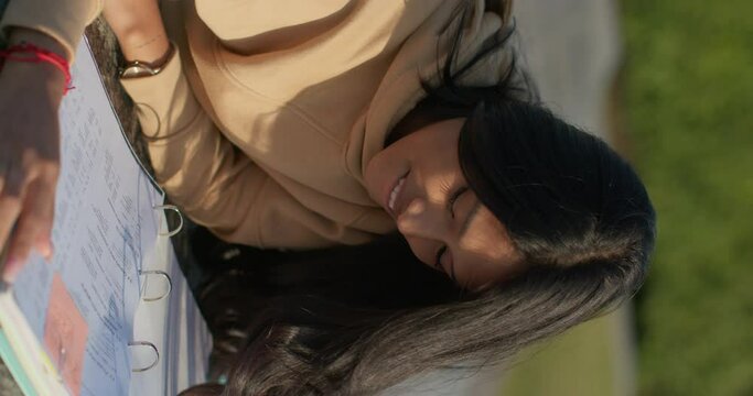 Young teenage Latina college student studying for an exam in an outdoor park with books, vertical shot