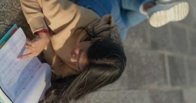 Young teenage Latina college student studying for an exam in an outdoor park with books, vertical shot