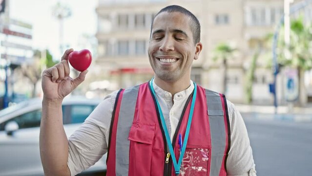 Young hispanic man volunteer smiling holding red heart for blood donation at street