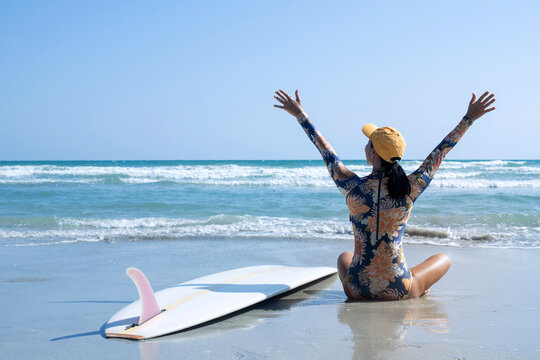 Female surfer sitting on beach in front of sea raise two hands up to sky feeling happy, outdoor activities, water sports activities concept