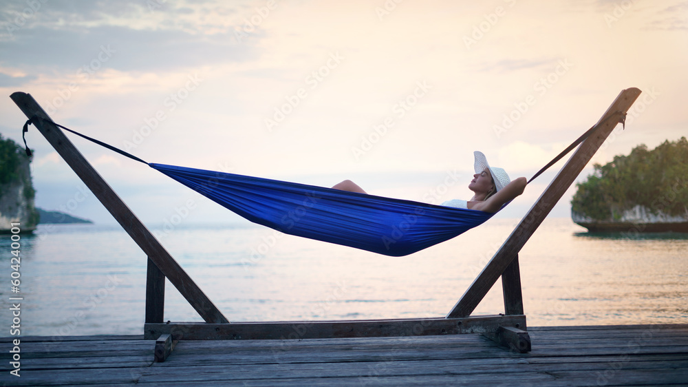 Wall mural Hammock, sunset ocean and woman relax outdoor on a vacation, holiday or adventure in nature. Female person on tropical trip, travel and deck with sea, sky and water in summer for zen, peace and calm