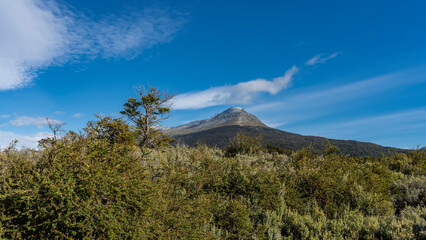 A mountain with a snowy peak on a background of blue sky and clouds. There is green vegetation in the foreground. Argentina. Ushuaia. Tierra del Fuego National Park