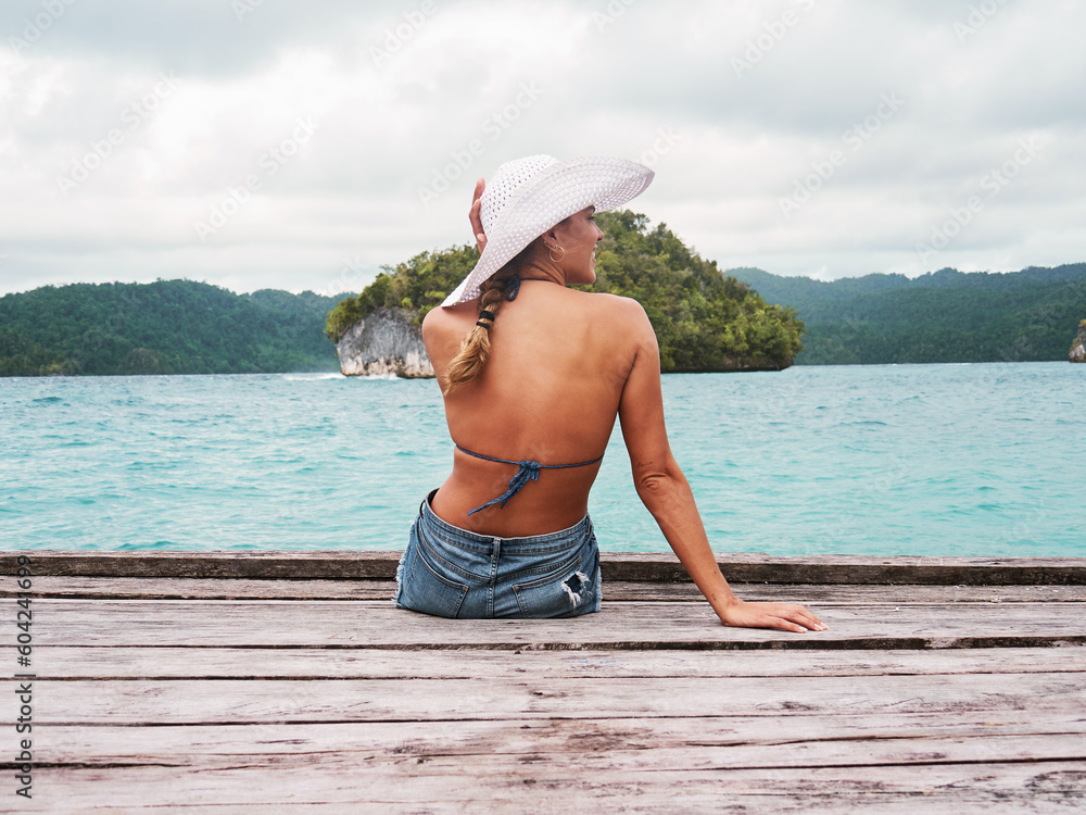 Wall mural Adventure, hat and woman on an island to relax while on tropical summer vacation or holiday. Travel, outdoor and back of female person sitting on wood pier by ocean water on weekend trip in paradise.