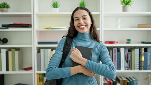 Young beautiful hispanic woman student standing holding book at university classroom