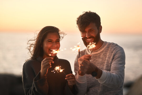 Sunset, beach and couple with fireworks for celebration, party and sparks on romantic date. Nature, love and happy man and woman with sparklers for honeymoon on holiday, weekend and vacation