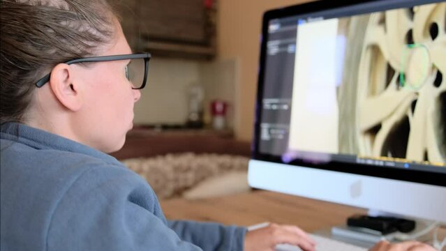 the camera shoots around a girl in glasses who sits at her desktop at home in the kitchen at the computer and works editing at photos, typing on the keyboard