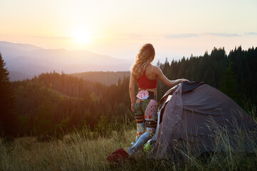 Woman tourist camping outdoors at sunset. Back view of slim woman near tent, admiring landscape in...