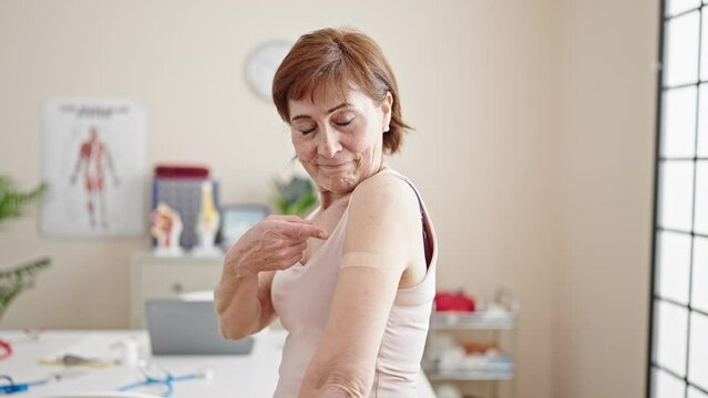 Mature hispanic woman doing thumbs up with band aid on arm at clinic