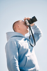 A man is holding a camera and looking through his lens against blue sky.