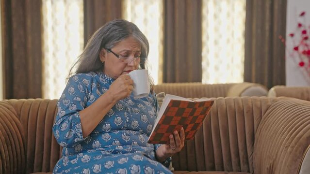 Tensed Indian aged woman reading book and drinking tea