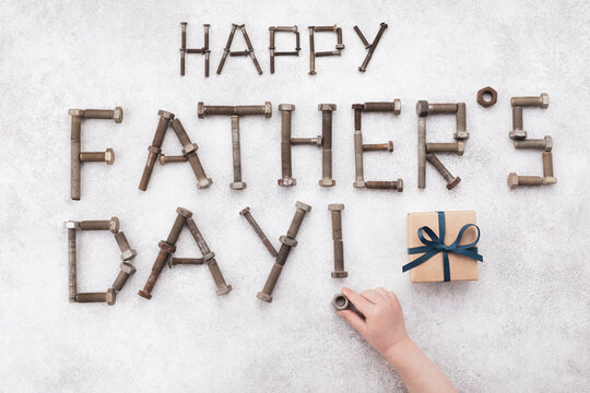 Happy fathers day greeting card from small child hand making lettering text from vintage screws and gift box top view on white stone background.