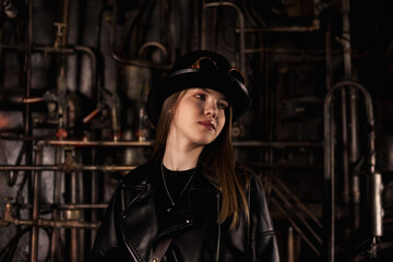Fototapeta na wymiar Pretty teen girl model in steampunk image in black leather jacket and top hat posing looking away. Teenage chic girl actress 12-13 year old in industrial room. Adventure style concept. Copy text space