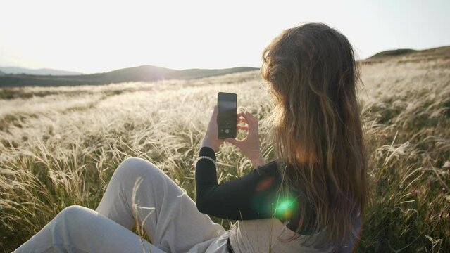 A modern woman in sun glare sits enjoying nature against the backdrop of a feather grass shining in the rays of a sunny sunset and the wind blows her hair. She takes pictures with her smartphone