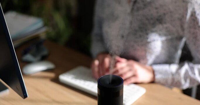 Modern air humidifier device during working hours. Manager using computer work on the Internet, enjoying aroma of aromatherapy steam from diffuser