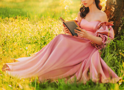 Fantasy woman sits under tree holding romantic book in hands reading novel smiling face, lips close-up. Pink long dress. Fairy girl in summer nature green grass magic sun light. art photo real people.