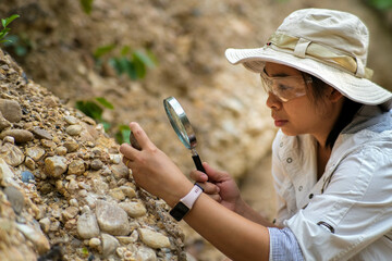 Female geologist using magnifying glass to examine and analyze rock, soil, sand in nature. Archaeologists explore the field. Environmental and ecology research. - obrazy, fototapety, plakaty