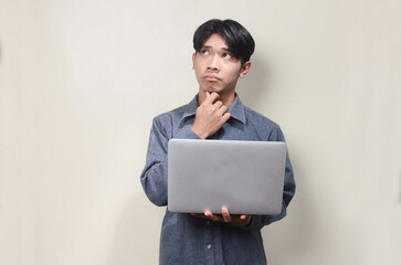 Asian young man thinking hard while carrying laptop on isolated background