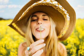 Beautiful young blonde cheerful woman, blowing dandelion seeds amidst a field of blooming yellow rapeseed flowers