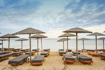 Wooden beach loungers with soft cushions and straw sun umbrellas right on the sea in a tourist resort in Greece against a cloudy blue sky, copy space