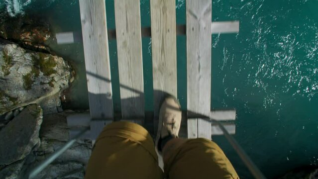 POV of man in hiking boots walk over flimsy wooden suspension bridge over wild mountain river. Epic hike adventure, or outdoor exploration concept. Shoes walk on wooden bridge