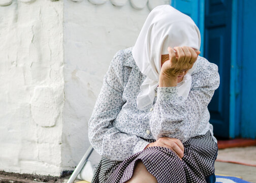 Upset Ukrainian senior woman sits near her house. Old age, loneliness. Russian aggression against Ukraine