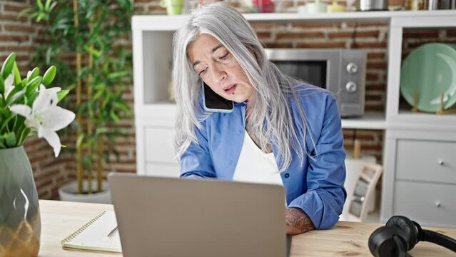 Middle age grey-haired woman using laptop talking on smartphone sitting on table at dinning room