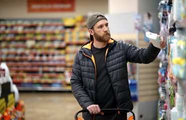 A bearded Caucasian man customer is interested in hygiene products at the supermarket - obrazy, fototapety, plakaty