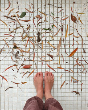 low section of woman standing barefoot on white tiled floor with scattered leaves
