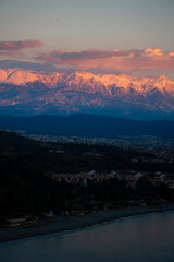 Snowy mountains in a pink sunset, with clouds, and a city at the foot that goes into the water